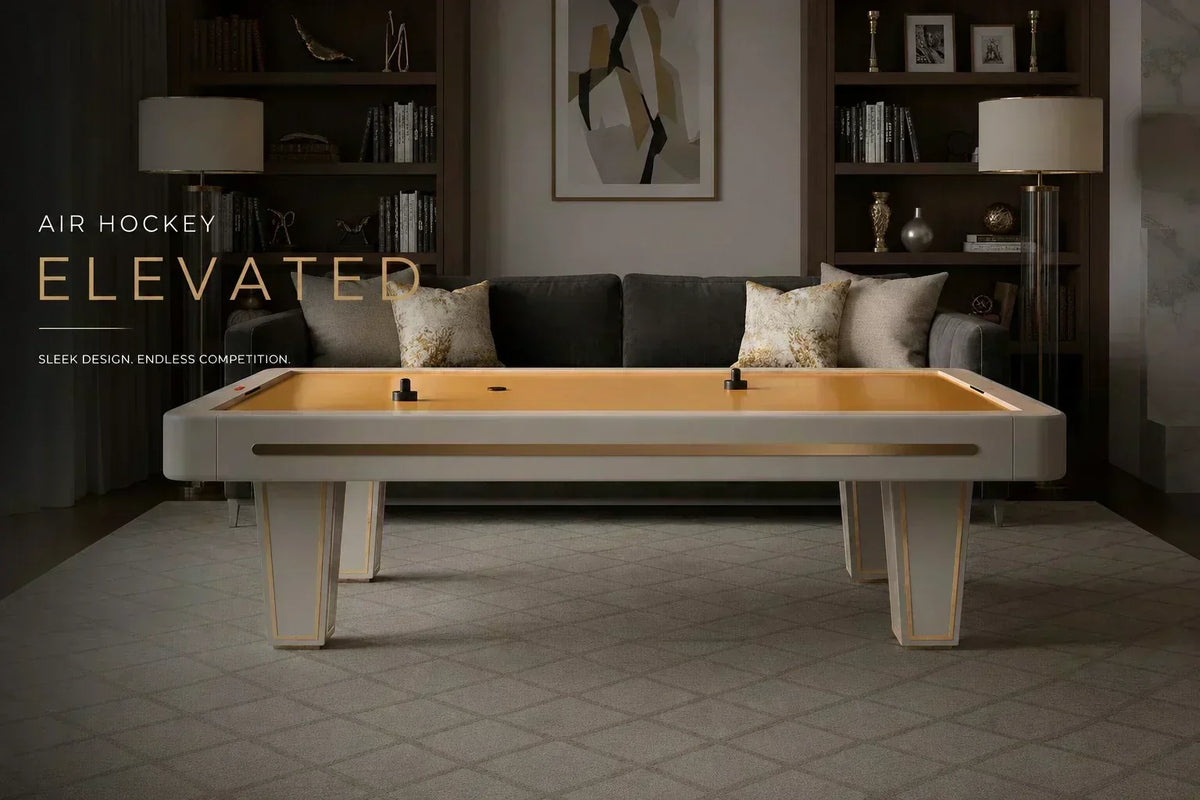 ARGMAC Vindsor air hockey table in cream with a golden playing surface, air hockey pucks and pushers on top, set in a dark, living room with a grey sofa and two gold-trimmed lamps.